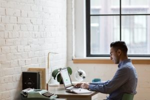 Side view of man working on laptop at desk in corner of loft space near window-min