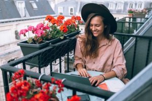 Smiling young woman sitting outside on balcony, working on laptop, surrounded by planters of red flowers in a picturesque European city