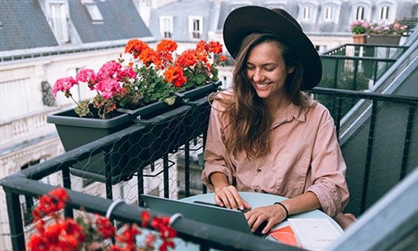 Smiling young woman sitting outside on balcony, working on laptop, surrounded by planters of red flowers in a picturesque European city