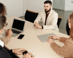 _View of new hire seated with hands placed on table surrounded by three high level execs in office space