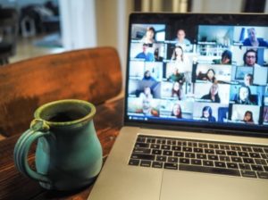 Close up of coffee cup beire laptop showing gallery view of multiple tiles of people in a video conference.