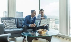 Man comfortably sitting on couch with feet on table in brightly lit corner office intently working on computer