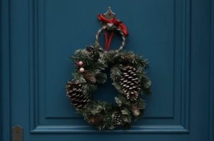 Close up dark teal door with holiday wreath made from pine cones hanging from brass knocker outside