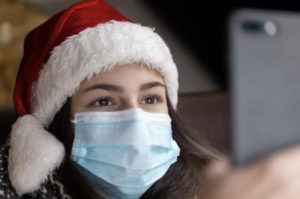 Close up of young woman wearing a santa hat and face mask, holding up smartphone to take a picture