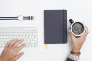 Overhead view of stylistically place objects hand on keyboard laid out watch, notebook and hand holding coffee on white surface