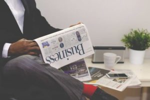 View of well-dressed man in suit sitting cross-legged reading business section of newspaper with table, plant and rest of newspaper in background