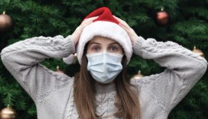 Young woman wearing a santa hat and face mask appearing surprised, with arms raised and placed on her head, standing in front of a large holiday tree