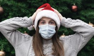 Mulher jovem usando um chapéu de Papai Noel e uma máscara facial parecendo surpresa, com os braços levantados e colocados na cabeça, em frente a uma grande árvore de feriado