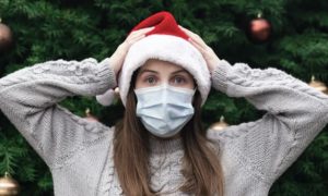 Young woman wearing a santa hat and face mask appearing surprised, with arms raised and placed on her head, standing in front of a large holiday tree-tile