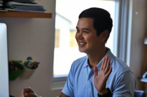 Side view of a smiling young man waving at his desktop, seated at desk in home office