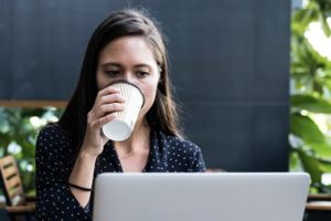 Woman diligently working at her laptop in outdoor cafe while sneaking in a sip of her takeaway coffee without removing her eyes from the screen