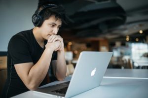 Focused-looking young man seated at desk in communal space with headphone, engaged with open laptop