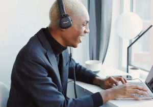 Happy, well-dressed man seated at modern-looking desk with headphones laughing and typing on open laptop near sunlit window