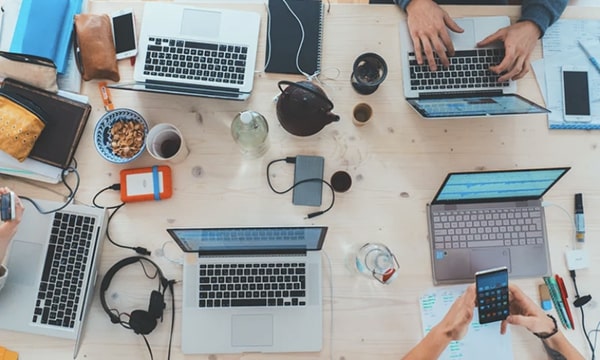 Overhead view of multiple laptops open with people working on them in a busy “war room” type of desk setting-tile