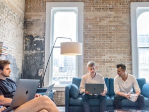 Wide view of team of two guys in front and two guys on the side working on couch with open laptops, in exposed brick, lofty office with high ceilings