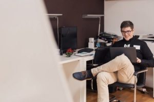 Young man facing away from desk with legs crossed, and open laptop on lap, smiling and interacting with screen