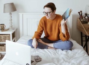 Young woman sitting cross-legged on bed at home with open laptop, looking at screen holding a closed book in her hand