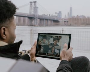 Over the shoulder view of man sitting outside near water by bridge, holding up a tablet with gallery view of faces in a video conference