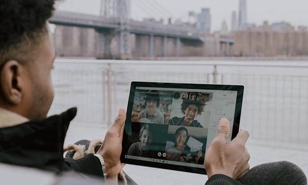 Over the shoulder view of man sitting outside near water by bridge, holding up a tablet with gallery view of faces in a video conference-tile