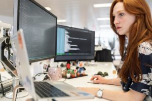 Side view of a female developer, focused and working on two desktop screens and a laptop in a brightly-lit office setting