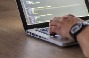 Up close view of a hand with a watch typing on a laptop with a screen full of coding