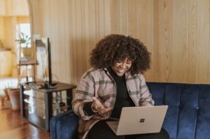 Wide angle view of happy woman sitting on couch at home smiling and looking at laptop with hand outreached and gesticulating