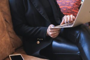 Close up of lower half of woman stylishly dressed, sitting on couch working on laptop with mobile device beside her