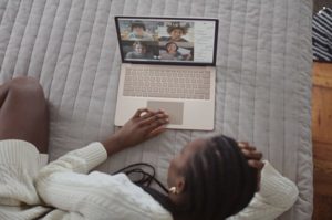 Overhead view of woman reclined on bed at home engaged on laptop in a video chat with four participants in Gallery View