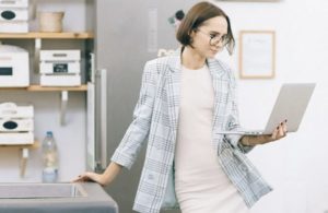 Business casual woman leaning on one arm against work table while holding an open laptop and reading from it with the other arm