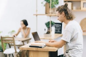 Group of three people crowded around a laptop on work desk in sunny workspace, chatting and writing in notebook