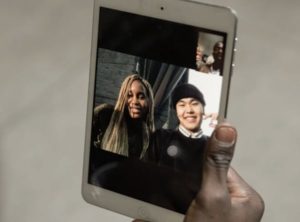 Hand holding up tablet device of a young man and woman smiling with a small picture-in-picture of a man and colleagues