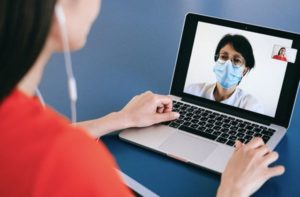 Over the shoulder view of a woman video chatting on laptop to a doctor in face mask