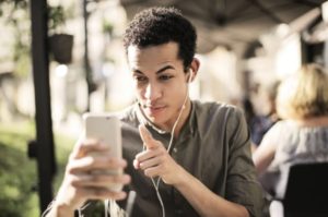 Young man outdoors on patio wearing headphones and interacting with device, pointing finger, and making a funny, serious face