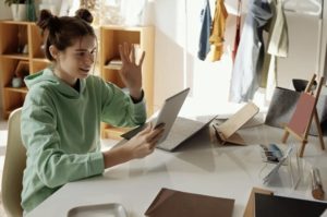 Young student working at desk in bedroom, smiling and interacting with tablet, holding up her hand waving