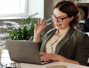 Young woman seated as desk in office wearing business attire smiling and introducing herself online via her laptop