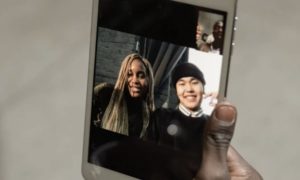 tile Hand holding up tablet device of a young man and woman smiling with a small picture-in-picture of a man and colleagues
