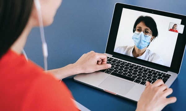 Over the shoulder view of a woman video chatting on laptop to a doctor in face mask-tile