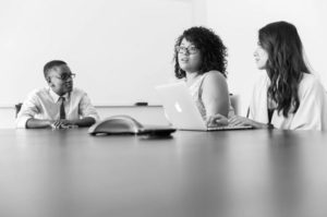 Black and white photo of table in foreground and team of three in midground, chatting working on laptop and engaging in conference call