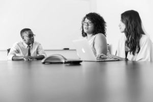 Fotografía en blanco y negro de la mesa en primer plano y un equipo de tres en el medio, charlando trabajando en una computadora portátil y participando en una llamada de conferencia