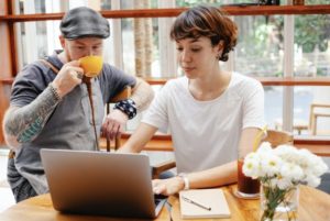 Stylish man drinking coffee looking at laptop while woman taps on keyboard and shows him content on screen, seated at table with white flowers nearby beside window