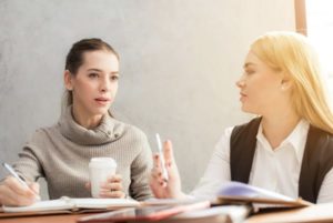 Two women chatting at table with open books. One is looking in the distance to the right of the camera while the other is chatting to her