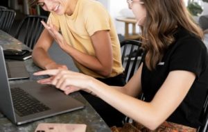 Two women engaged in discussion, laughing and pointing at their laptops on desk in communal work space