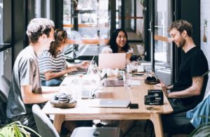 View of four happy team members seated at a long desk table working on laptops, laughing and chatting in brightly lit communal work space