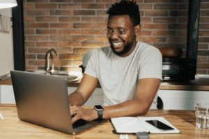 View of smiling man seated diagonally at table working on laptop in communal office space kitchenette with opened notebook and device