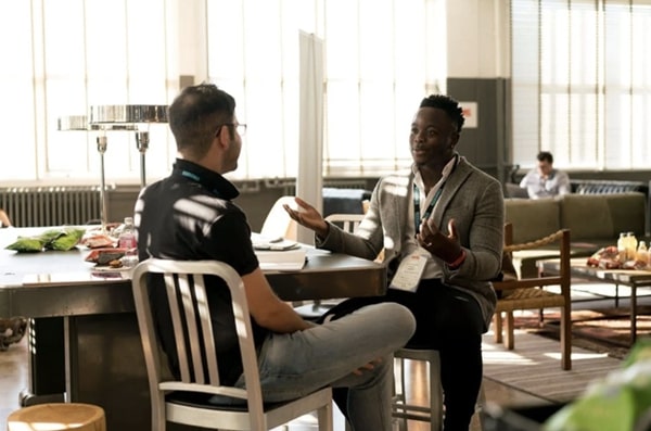 View of two men sitting at corner of table in brightly lit, stylish communal workspace engaged in lively conversation