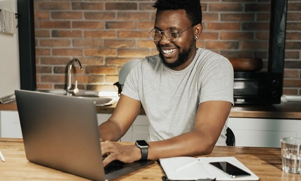 View of smiling man seated diagonally at table working on laptop in communal office space kitchenette with opened notebook and device-tile