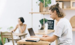 tile-Group of three people crowded around a laptop on work desk in sunny workspace, chatting and writing in notebook