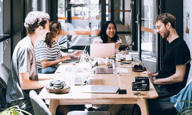 tile-View of four happy team members seated at a long desk table working on laptops, laughing and chatting in brightly lit communal work space