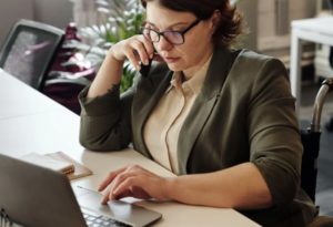 Close up view of business casual woman chatting on phone seated at table in front of laptop working away.