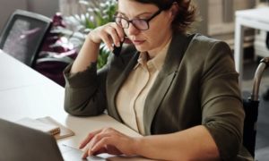 Close up view of business casual woman chatting on tile-phone seated at table in front of laptop working away